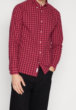Homme portant une chemise à carreaux rouge boutonnée avec un pantalon noir, debout avec une main dans la poche sur un fond uni.