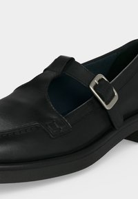 Chaussure Mary Jane en cuir noir avec une bride à boucle argentée et un bout arrondi, présentée sur un fond blanc uni.