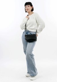 Donna che indossa un maglione di lana bianco e jeans azzurri chiari con scarpe da ginnastica bianche, che porta una borsa a tracolla nera, in piedi su uno sfondo bianco.