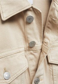 Chaqueta de mezclilla beige con botones plateados, cuello puntiagudo y detalles en los bolsillos. La textura del tejido es suave con un aspecto ligeramente desgastado.