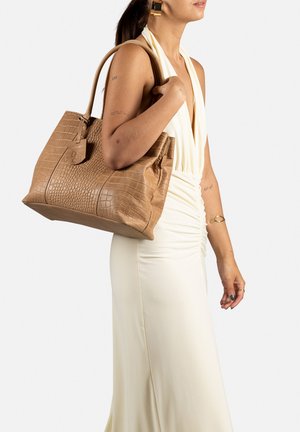 Tan krokodillenleer handtas met bovenhandvaten, voorzien van een ritssluiting en een voorlabel, vastgehouden door een persoon in een witte jurk.