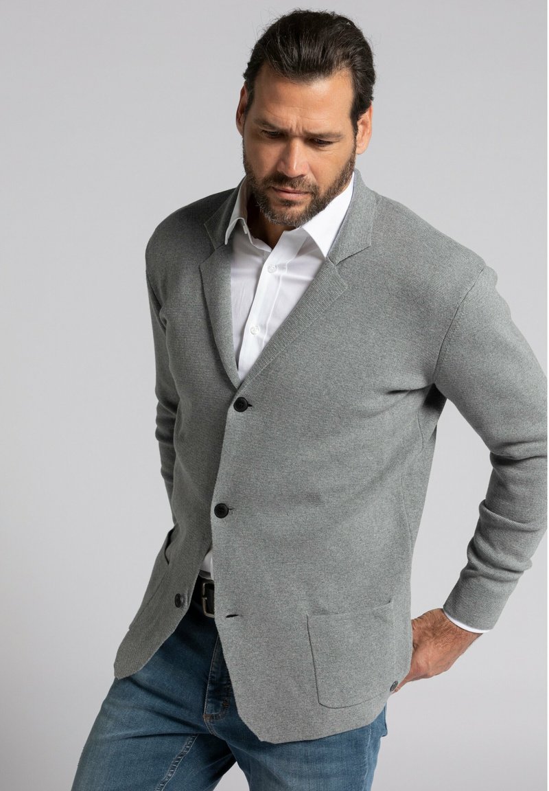 Cardigan gris hecho de tejido de punto, con un cuello de muesca, cierre de un solo botón y dos bolsillos frontales. Llevado sobre una camisa blanca.