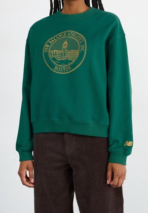 Person, die einen grünen Sweatshirt mit einem goldenen New Balance Athletics Boston-Logo und braune Cordhosen trägt, vor einem einfarbigen Hintergrund steht.