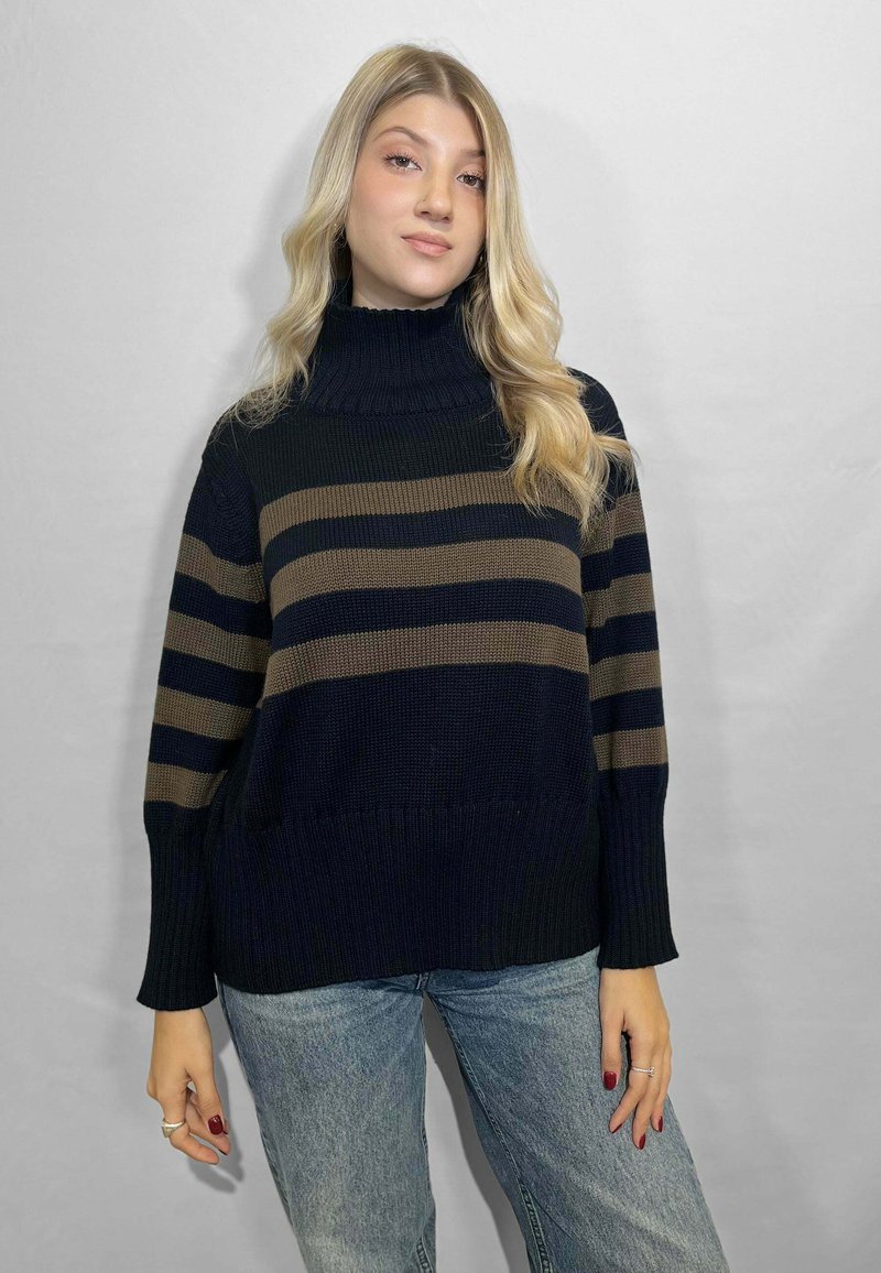 Giovane donna con lunghi capelli biondi indossa un maglione nero a collo alto con righe marroni e jeans azzurri, in piedi di fronte a uno sfondo semplice.