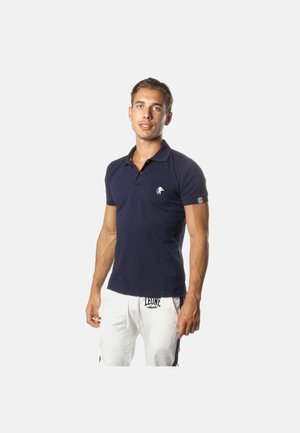 Uomo che indossa una polo blu navy con piccolo logo bianco sul petto e pantaloni bianchi con coulisse e scritta nera sul davanti, in piedi davanti a uno sfondo bianco.