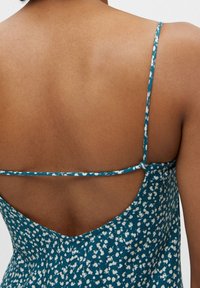 Mujer vistiendo un vestido verde azulado con estampado floral blanco y tirantes finos que se cruzan en una espalda baja abierta.