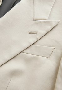 Gros plan du revers d'une veste de costume beige montrant la boutonnière et la poche poitrine avec la texture fine du tissu en laine.