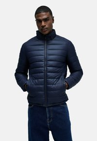 Granatowa pikowana kurtka z wysokim kołnierzem, zamek błyskawiczny i dwie kieszenie z przodu; ma poziome pikowanie.