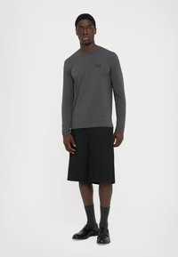 Man standing wearing a dark gray long-sleeve shirt, black knee-length shorts, dark socks, and black leather shoes against a white background.