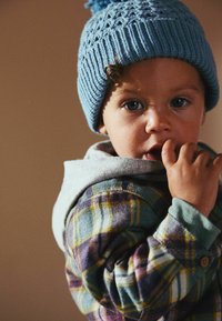 L'enfant porte un bonnet en tricot bleu clair avec un pompon, un sweat à capuche gris et une chemise à carreaux dans diverses couleurs, y compris le violet, le jaune et le bleu.