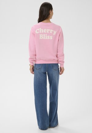 Kobieta stojąca tyłem do kamery, ubrana w różową bluzę z napisem „Cherry Bliss” i niebieskie dżinsy na jednolitym tle.