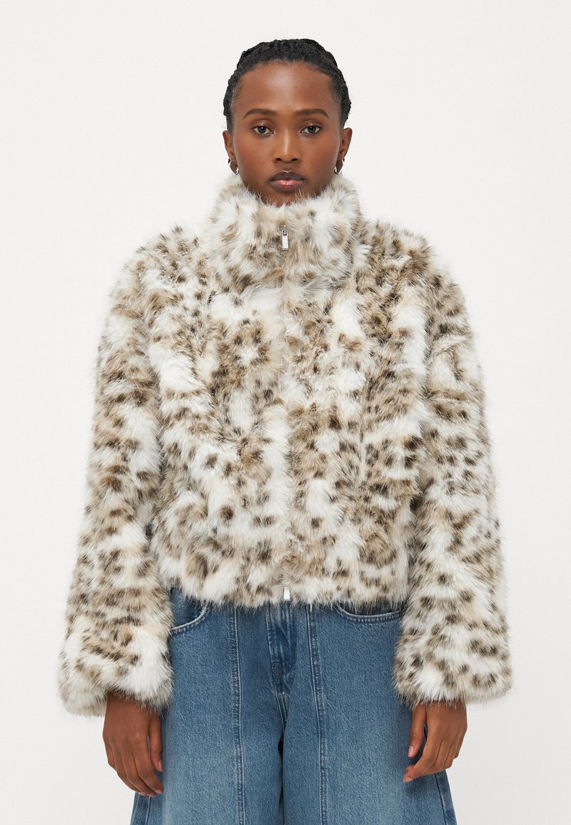 Faux fur-jakke med høy krage, med et flekkete mønster i brunt og hvitt. Den har lange ermer og en frontlynlås.