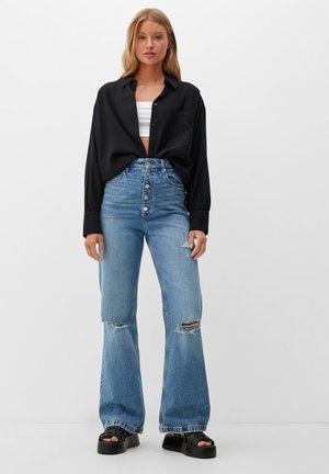 Hoogwaardige blauwe denim jeans met een flared pijpen, voorzien van een knoopsluiting, slijtage en een donker hemd met kraag dat over een crop top is gedragen.