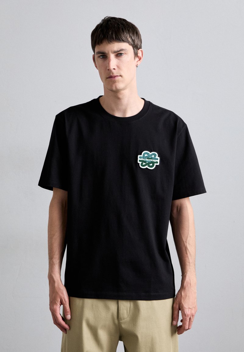 Black cotton t-shirt with short sleeves. Features a green embroidered patch on the left chest area. Simple design with a crew neckline.