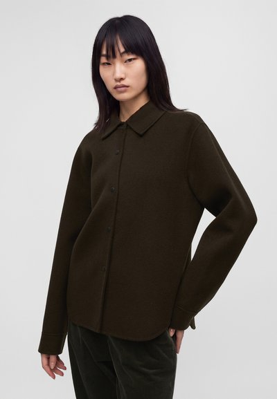 Femme aux cheveux noirs raides portant une veste en laine boutonnée de couleur olive foncé et un pantalon sombre sur un fond clair uni.