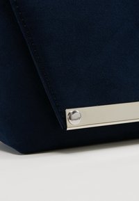 Kabelka psaníčko z tmavě modré semišové kůže s kovovým pruhem a detaily ve tvaru kruhových nýtů. Design je elegantní s čistými liniemi a texturovaným povrchem.