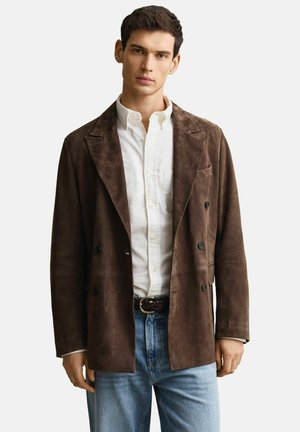 Junger Mann trägt eine braune, doppelt geknöpfte Wildlederjacke, weißes Hemd mit Knöpfen, geflochtener Gürtel und hellblaue Jeans.