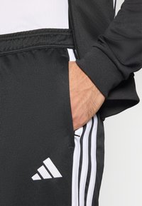 Pantalon de sport noir avec des bandes blanches sur les côtés, doté d'une taille élastique et d'un logo visible sur le bas de la jambe.