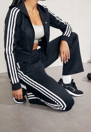 Osoba klęcząca w czarnym dresie marki Adidas z białymi paskami, jasnoszarym sportowym biustonoszu, białych skarpetkach i czarnych butach sportowych.