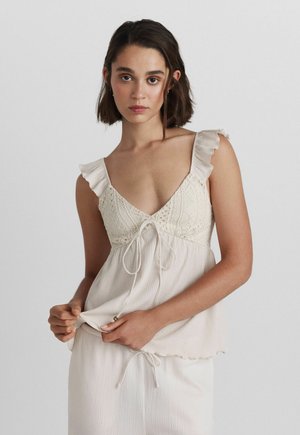 Jonge vrouw met kort donker haar die een mouwloze crèmekleurige top draagt met gerimpelde bandjes en een trekkoorddetail op de borst.