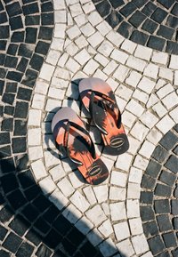 Paire de tongs Havaianas noires et orange avec motif de palmier sur un trottoir circulaire en carreaux noir et blanc.