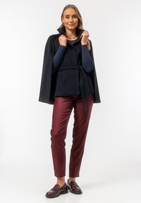 Granatowa bluzka w stylu peleryny z przewiązaniem w talii, zestawiona z burgundowymi spodniami slim fit i ciemnymi loafersami. Gładki materiał i zdefiniowany krój.