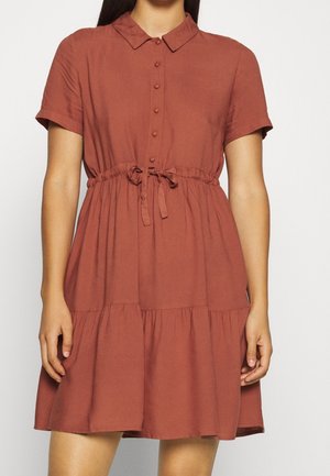 Femme portant une robe rouille à manches courtes, boutonnée, avec col et taille nouée, debout devant un fond clair uni.