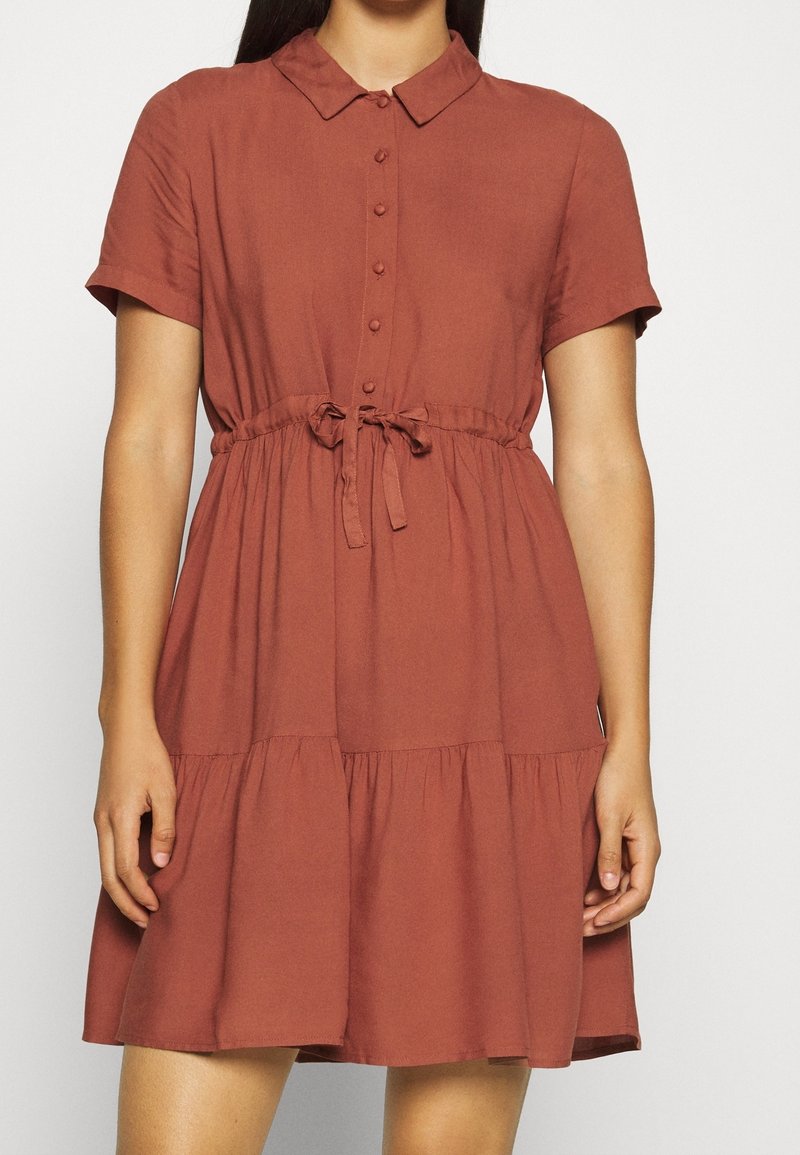Femme portant une robe rouille à manches courtes, boutonnée, avec col et taille nouée, debout devant un fond clair uni.