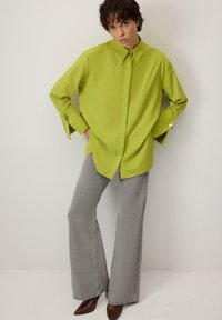 Laimerohelise tooniga ülepaistuv särk terava krae ja lõhestatud mansettidega, kombineeritud helehallide lõhenevate pükstega ja pruunide teravate ninadega saapadega.