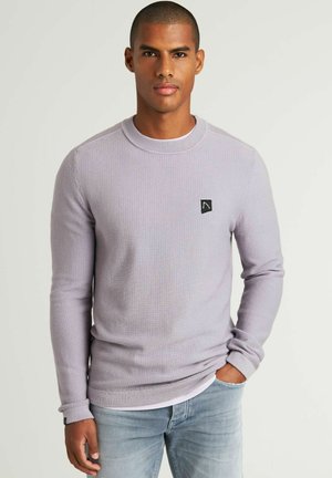 Suéter de punto texturizado de color morado claro, con mangas largas y cuello redondo, que presenta un pequeño logo negro en el lado izquierdo del pecho. Combinado con jeans de color azul claro.