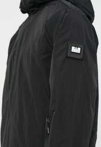 Sort hættejakke lavet af letvægtsmateriale, med en lynlåslomme og et mærket patch på venstre arm. Glat tekstur.