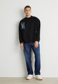Zwarte sweatshirt met een grafische zak; relaxed fit. Gecombineerd met blauwe jeans en witte sneakers met oranje zolen. Glad textuur van de stof.