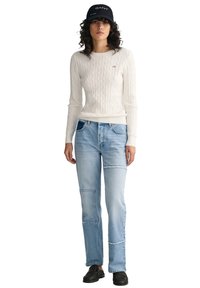 Weißer Kabelstrickpullover kombiniert mit hellblauen, geflickten Denim-Jeans. Das Modell trägt eine marineblaue Mütze und schwarze, geflochtene Slip-On-Schuhe.