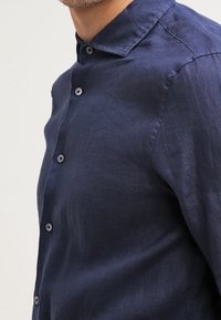 Marinblå linnekjorta med liten krage, knäppning fram och strukturerad tyg. Har kontrasterande knappar längs knäppningen.