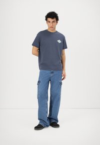 Camiseta de manga corta azul marino con un logo en forma de diamante de color azul claro en el pecho, combinada con unos pantalones cargo de ajuste holgado en azul claro y zapatos negros.