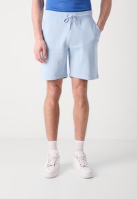 Shorts de sport bleu clair avec une taille élastique, cordon de serrage et poches latérales. Associés à des baskets blanches et des chaussettes de longueur mi-mollet.