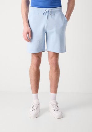 Hombre con pantalones cortos de cordón azul claro, calcetines blancos, zapatillas blancas y camisa azul, de pie con las manos en los bolsillos.