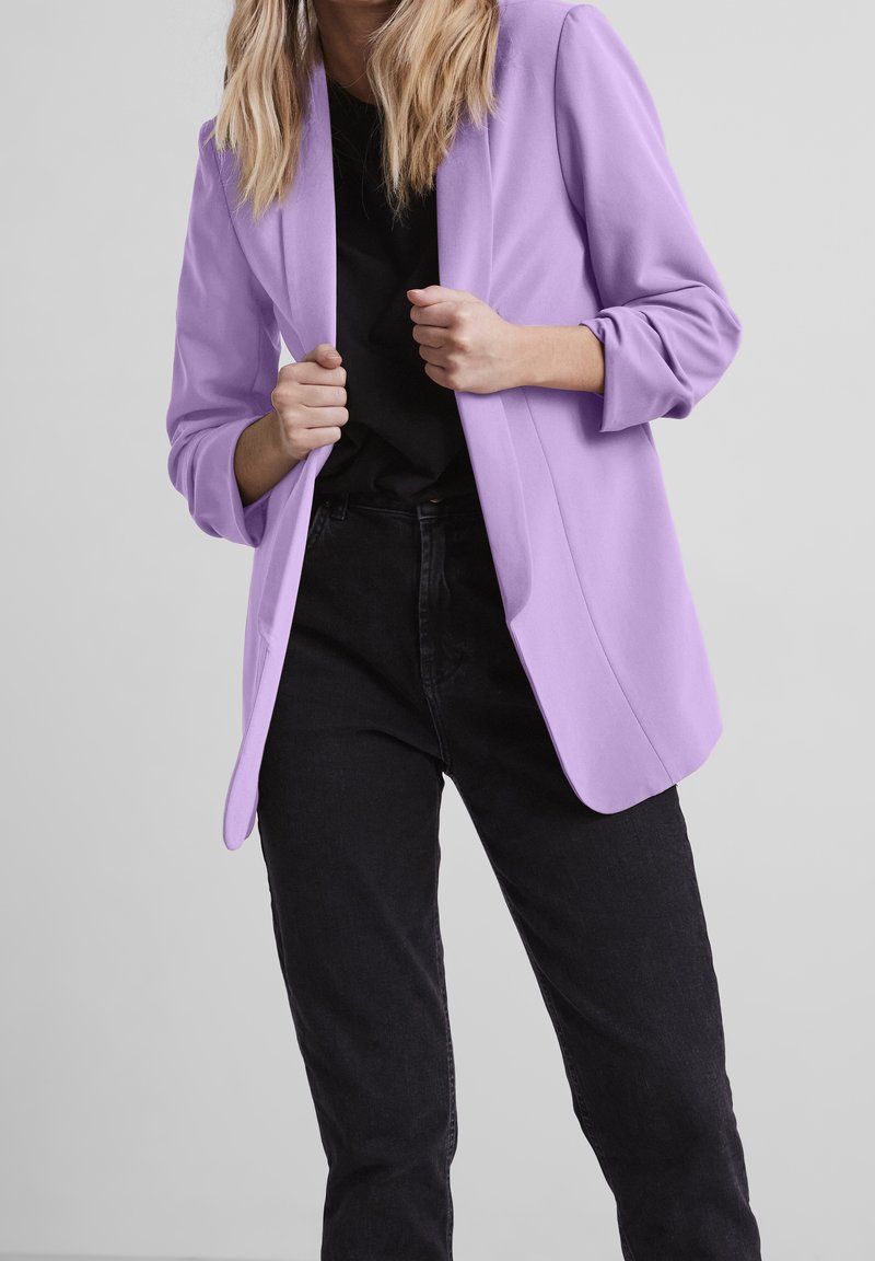 Femme portant un blazer lilas par-dessus une chemise noire et un pantalon noir, tenant les revers du blazer avec les deux mains sur un fond neutre.