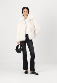 Giacca bianca in pelliccia sintetica con colletto ampio, abbinata a un top bianco aderente e jeans neri a zampa d'elefante. Tiene una borsa nera in pelle.