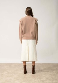 Beige gebreide trui met ribbelstructuur, gecombineerd met een witte rok van gemiddelde lengte met een centrale naad. Bruine enkellaarzen met blokhakken.