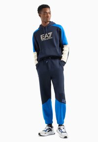 Sudadera con capucha azul marino con mangas en azul y crema, con el logo EA7. Combinada con pantalones azul marino con detalles en azul. Lleva zapatillas blancas.