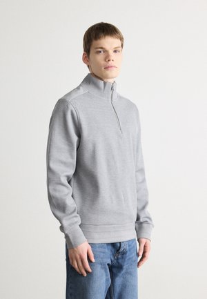 Sweatshirt gris à zip quart en tissu texturé, avec des poignets et un ourlet côtelés. Porté avec un jean bleu, debout devant un fond uni.