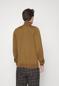 Man in een bruin coltrui en donkere geruite broek, staand met de rug naar de camera tegen een effen lichte achtergrond.