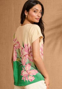 T-shirt a maniche corte di colore crema con motivo floreale, caratterizzata da fiori di iris rosa e dettagli verdi, realizzata in tessuto morbido con una vestibilità comoda e scollo rotondo.