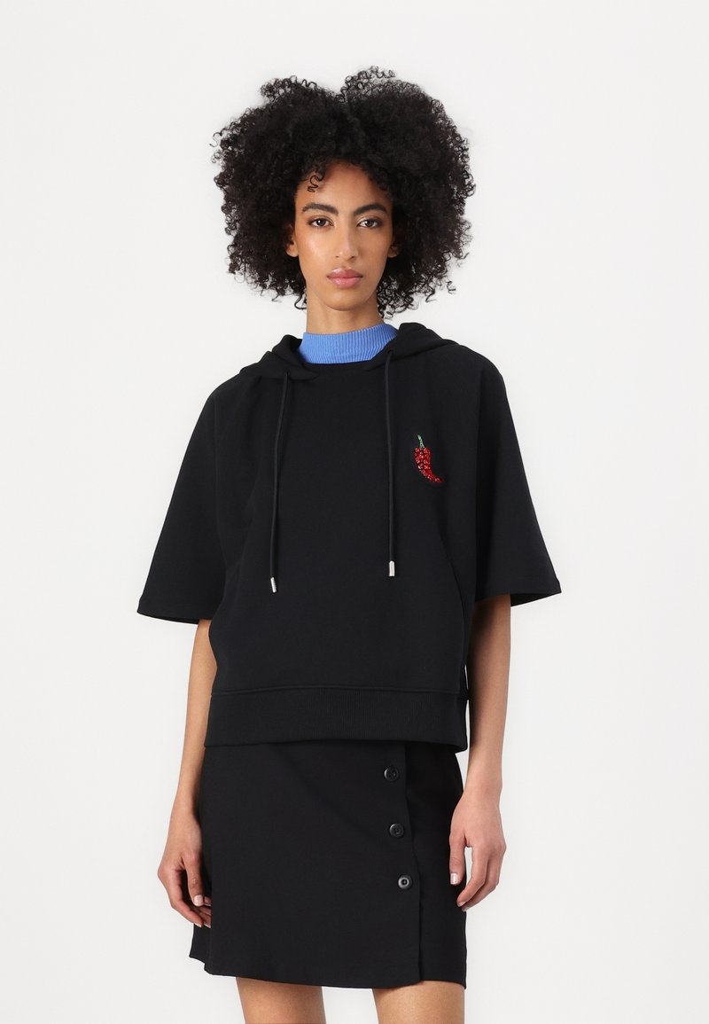 Sudadera con capucha negra de manga corta y cordones plateados, con un bordado de un pimiento rojo. Combinada con una falda negra de botones.