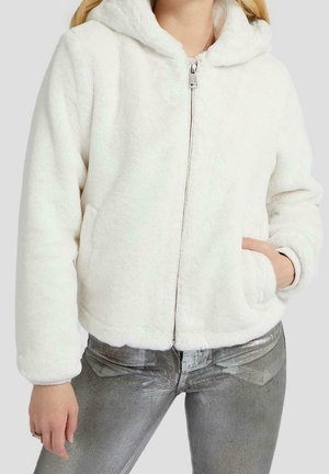 Femme portant une veste à capuche blanche et duveteuse avec une fermeture éclair et des poches, ainsi qu'un pantalon argenté métallique, debout avec une main dans la poche.