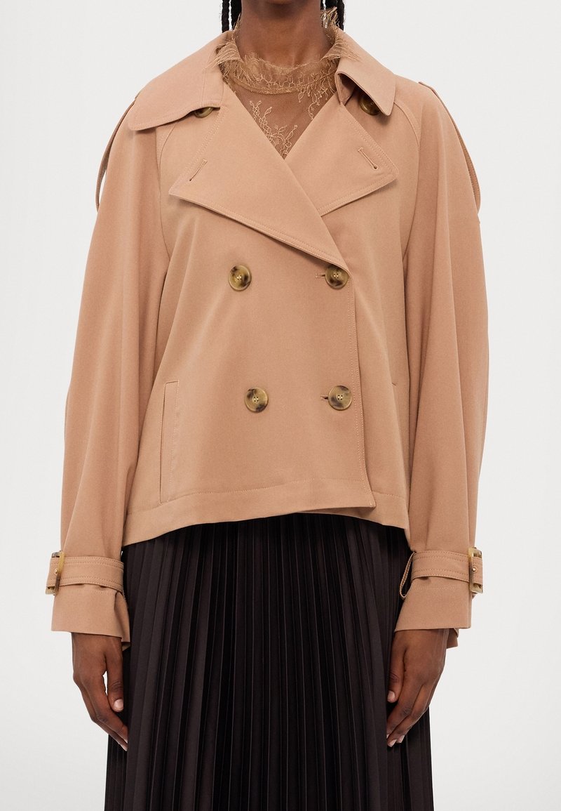 Femme portant un trench-coat court beige à double boutonnage avec de larges revers et une jupe plissée noire.