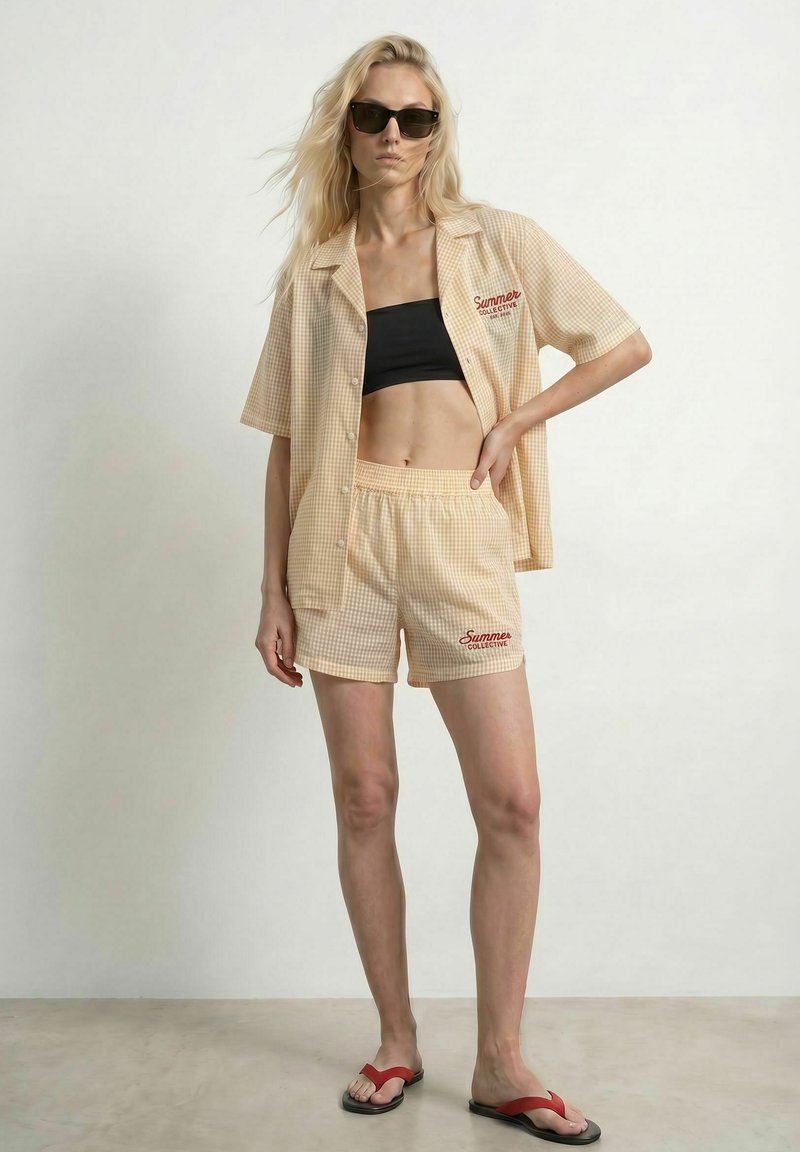 Femme portant des lunettes de soleil, chemise et short à carreaux beige avec le texte "Summer Collective", haut noir et sandales rouges, debout à l'intérieur.
