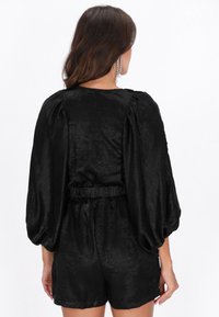 Fekete szatén overál kerek nyakkivágással, felfújt hosszú ujjakkal és elasztikus derékpánttal. Simas textúra enyhe fényességgel.