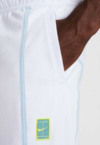 Bílé sportovní šortky s světle modrým pruhem po straně a nášivkou s logem Nike v zelené a žluté barvě na levé kapse.